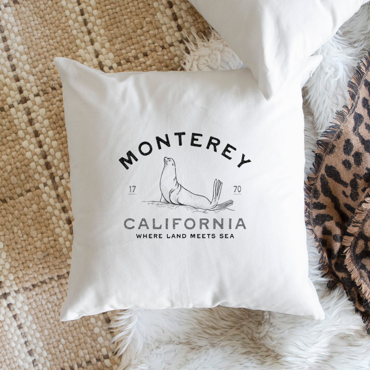 White pillow cover printed with a black ink sea lion with the words Monterey California, 1770, Where Land Meets Sea, on a woven chair.