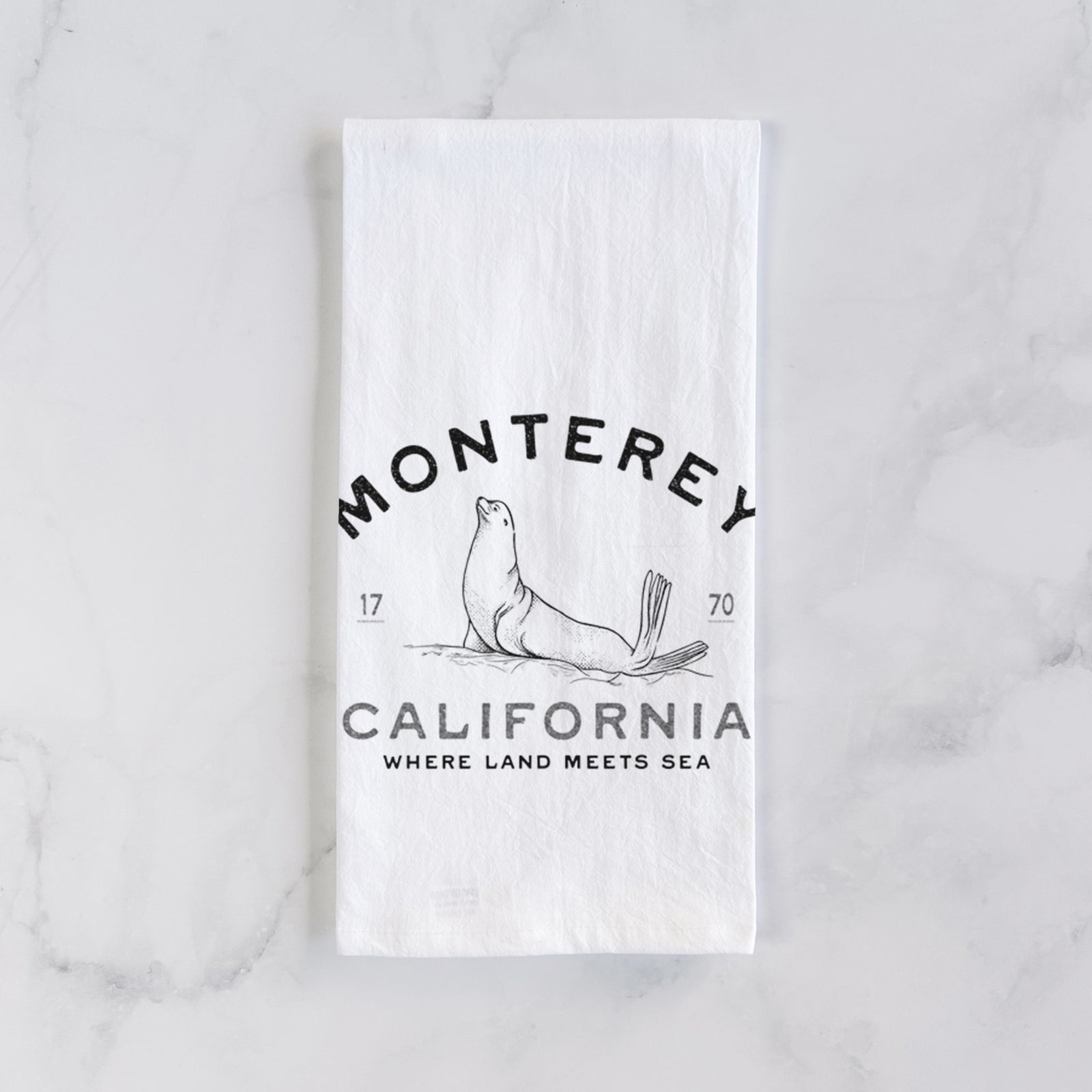 White cotton tea towel printed with a black ink sea lion with the words Monterey California, 1770, Where Land Meets Sea, on a marble counter beside a round wooden cutting board and greenery.