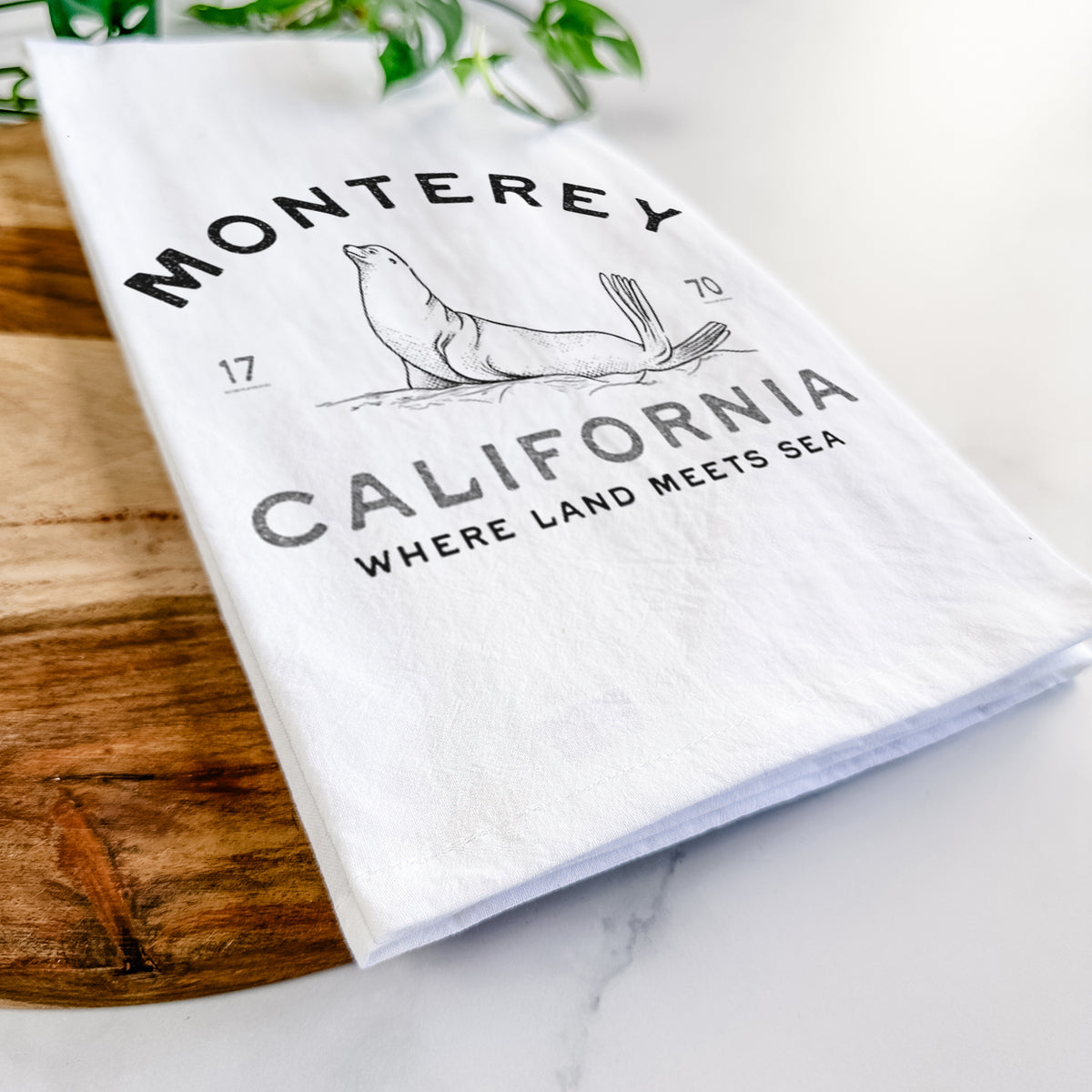 Close-up of a folded white tea towel on a wooden board, showing a black ink sea lion with the words Monterey California, 1770, Where Land Meets Sea.