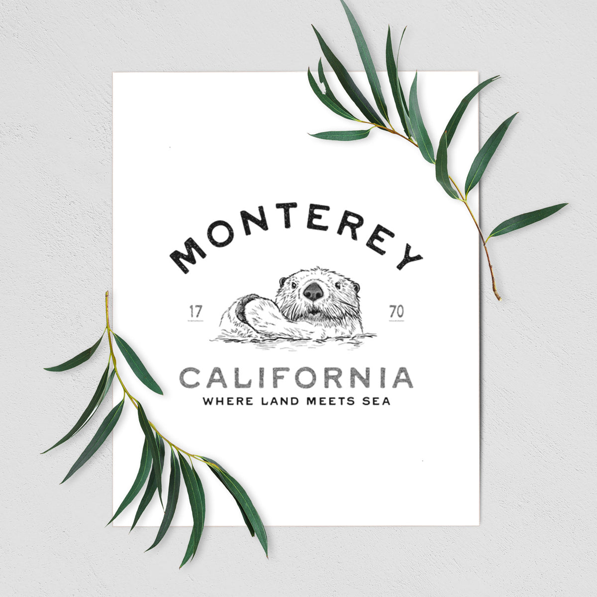 Art print of a black ink sea otter floating on its back, with the words Monterey California, 1770, Where Land Meets Sea, shown with eucalyptus leaves.