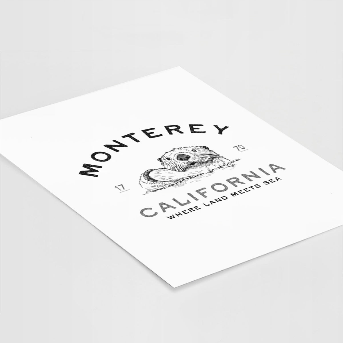Art print of a black ink sea otter floating on its back, with the words Monterey California, 1770, Where Land Meets Sea, lying at an angle on a white surface.