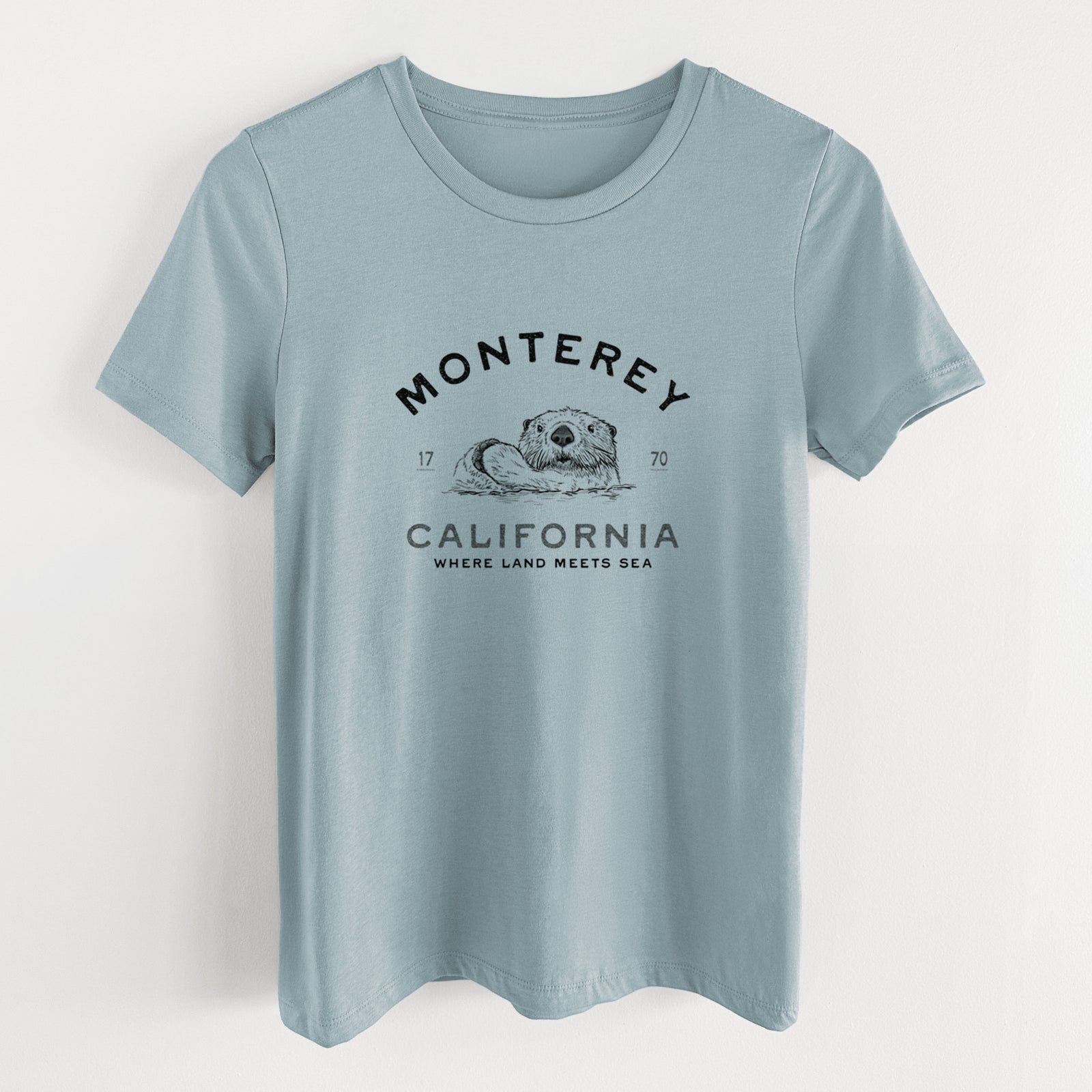 Vintage denim women's relaxed fit cotton t-shirt laid flat, printed with a black ink sea otter floating on its back, with the words Monterey California, 1770, Where Land Meets Sea.