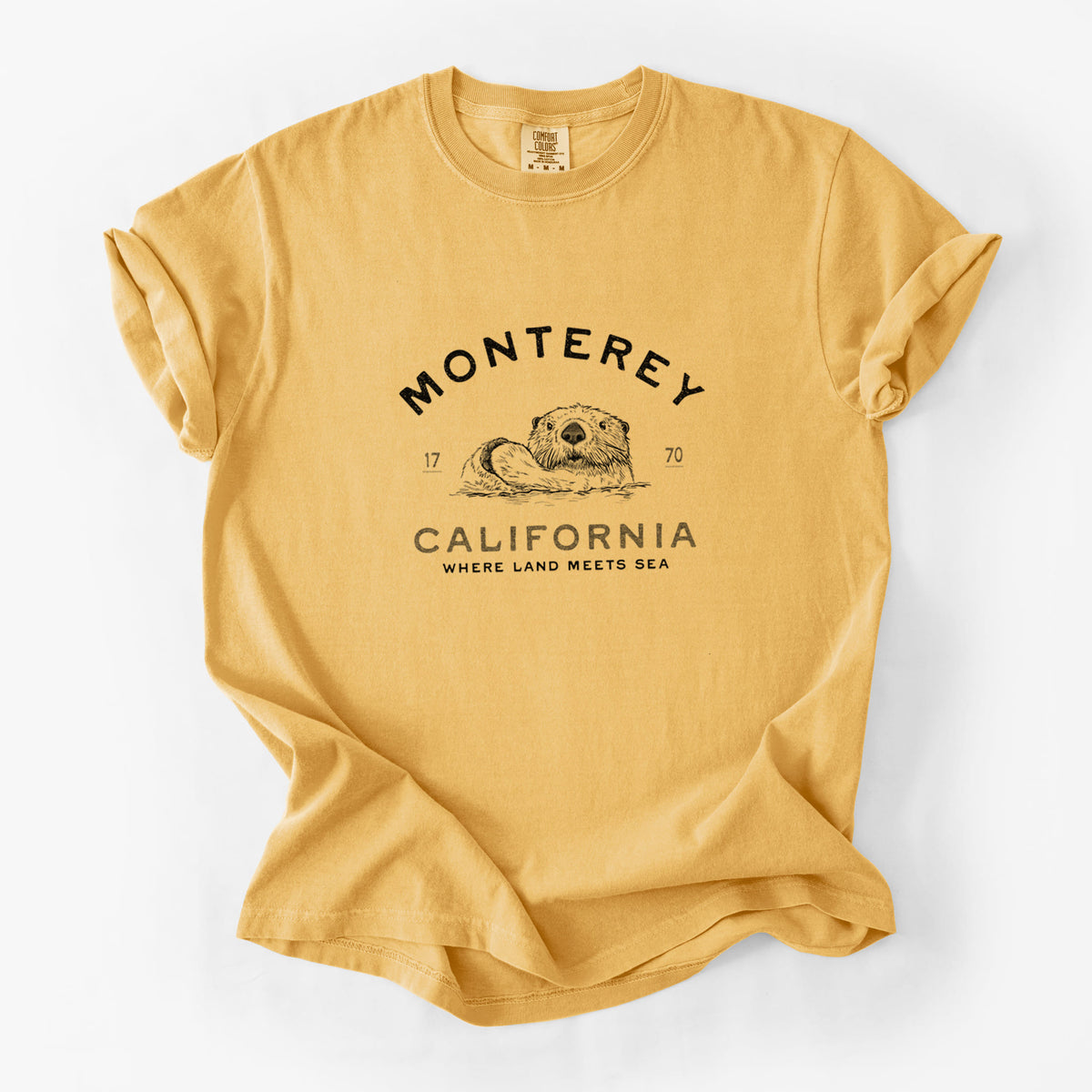 Golden wheat yellow Comfort Colors t-shirt laid flat, printed with a black ink sea otter floating on its back, with the words Monterey California, 1770, Where Land Meets Sea.