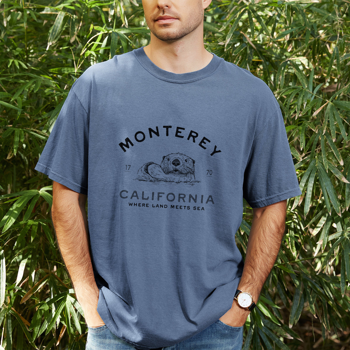 Man wearing a Comfort Colors t-shirt printed with a black ink sea otter floating on its back, with the words Monterey California, 1770, Where Land Meets Sea, outdoors.