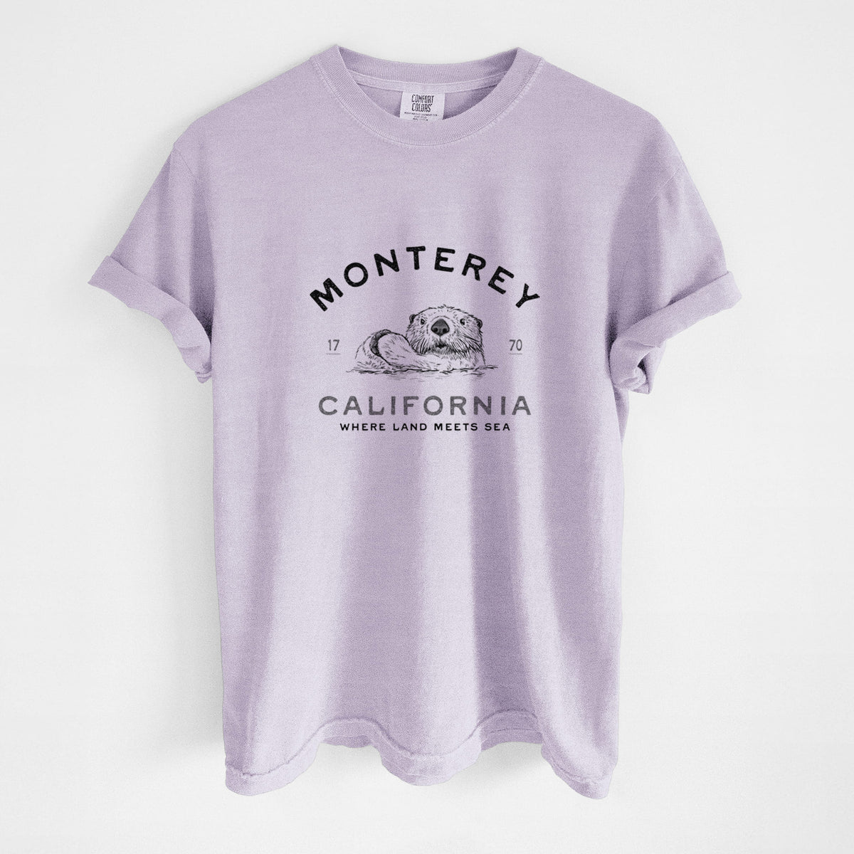 Orchid purple Comfort Colors t-shirt laid flat, printed with a black ink sea otter floating on its back, with the words Monterey California, 1770, Where Land Meets Sea.