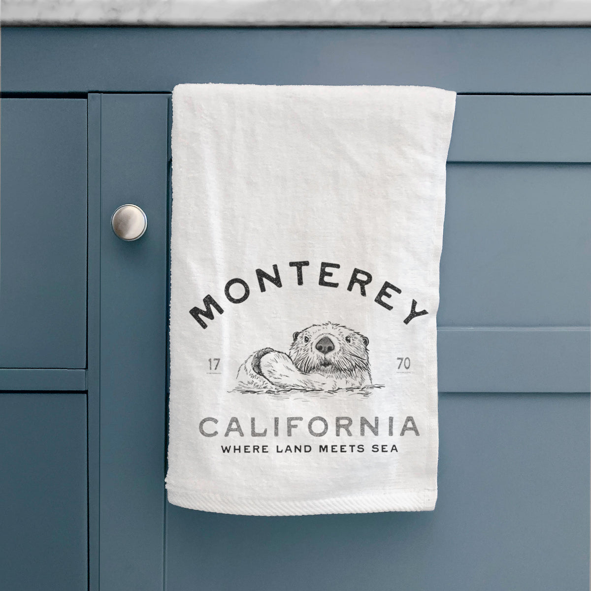 White hand towel printed with a black ink sea otter floating on its back, with the words Monterey California, 1770, Where Land Meets Sea, hanging on a cabinet door.
