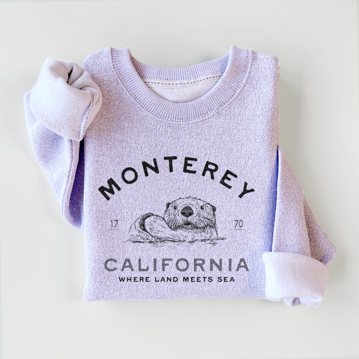 Ice purple knit sweatshirt printed with a black ink sea otter floating on its back, with the words Monterey California, 1770, Where Land Meets Sea, styled flat.