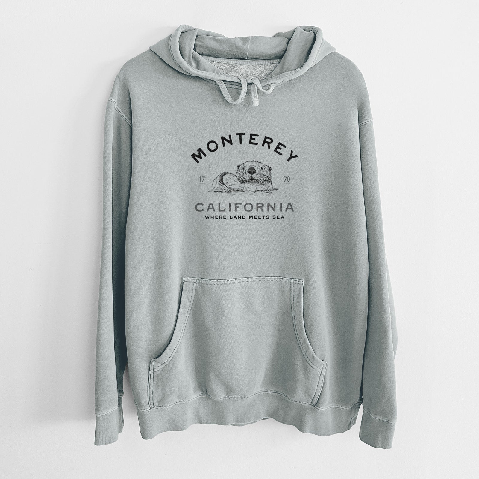 Sage green pigment dyed hoodie laid flat, printed with a black ink sea otter floating on its back, with the words Monterey California, 1770, Where Land Meets Sea.