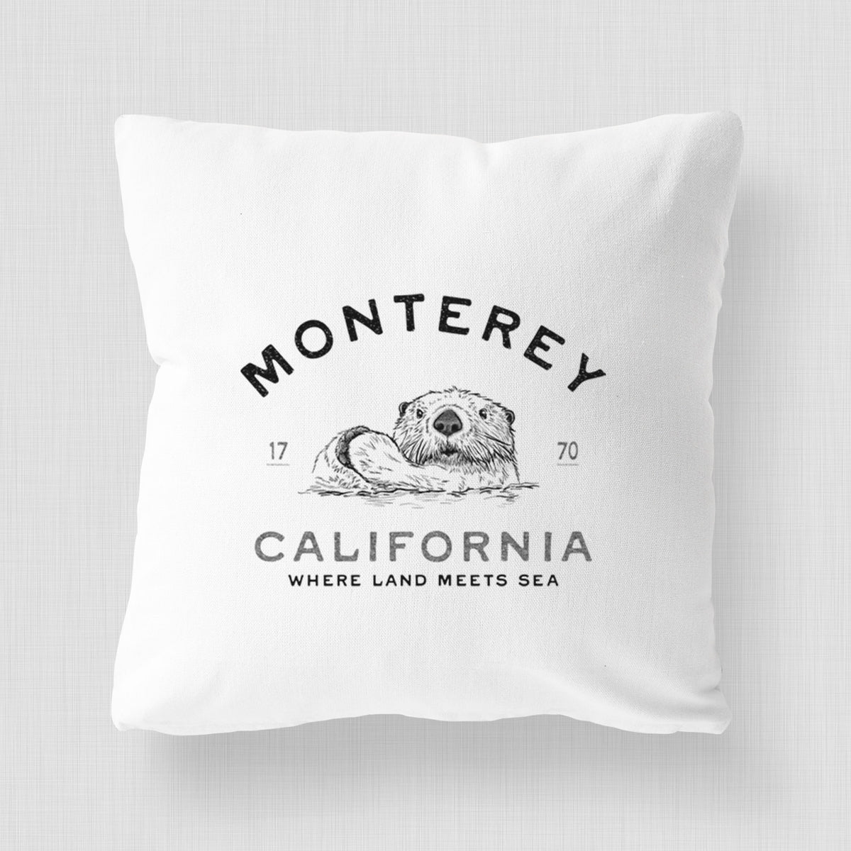 White pillow cover printed with a black ink sea otter floating on its back, with the words Monterey California, 1770, Where Land Meets Sea.
