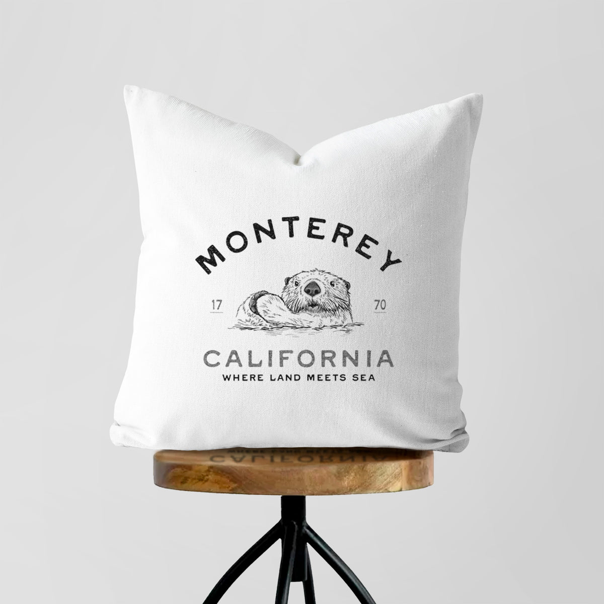 White pillow cover printed with a black ink sea otter floating on its back, with the words Monterey California, 1770, Where Land Meets Sea, on a wooden stool.