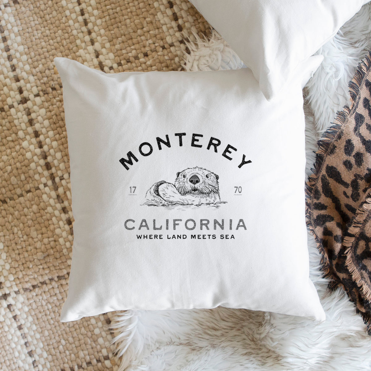 White pillow cover printed with a black ink sea otter floating on its back, with the words Monterey California, 1770, Where Land Meets Sea, on a woven chair.