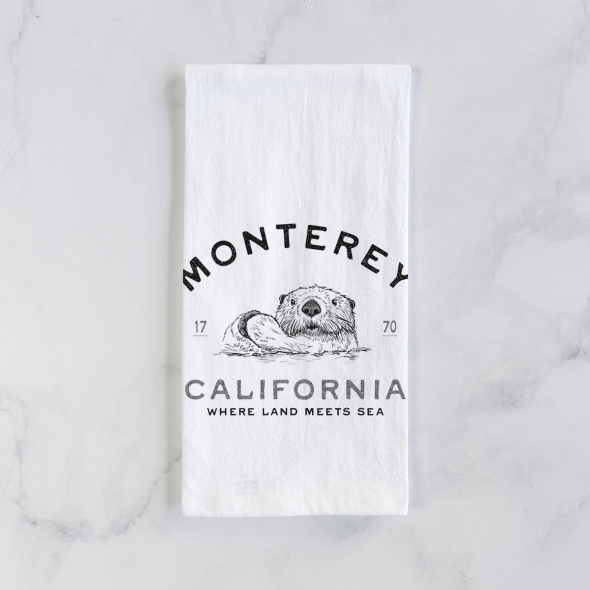 White cotton tea towel laid flat on marble, printed with a black ink sea otter floating on its back, with the words Monterey California, 1770, Where Land Meets Sea.
