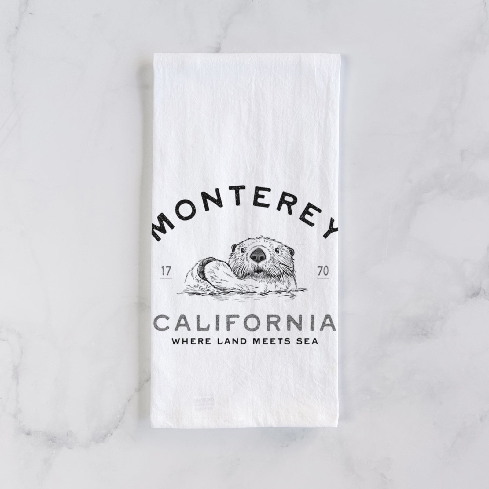 White cotton tea towel printed with a black ink sea otter floating on its back, with the words Monterey California, 1770, Where Land Meets Sea, on a marble counter beside a round wooden cutting board and greenery.