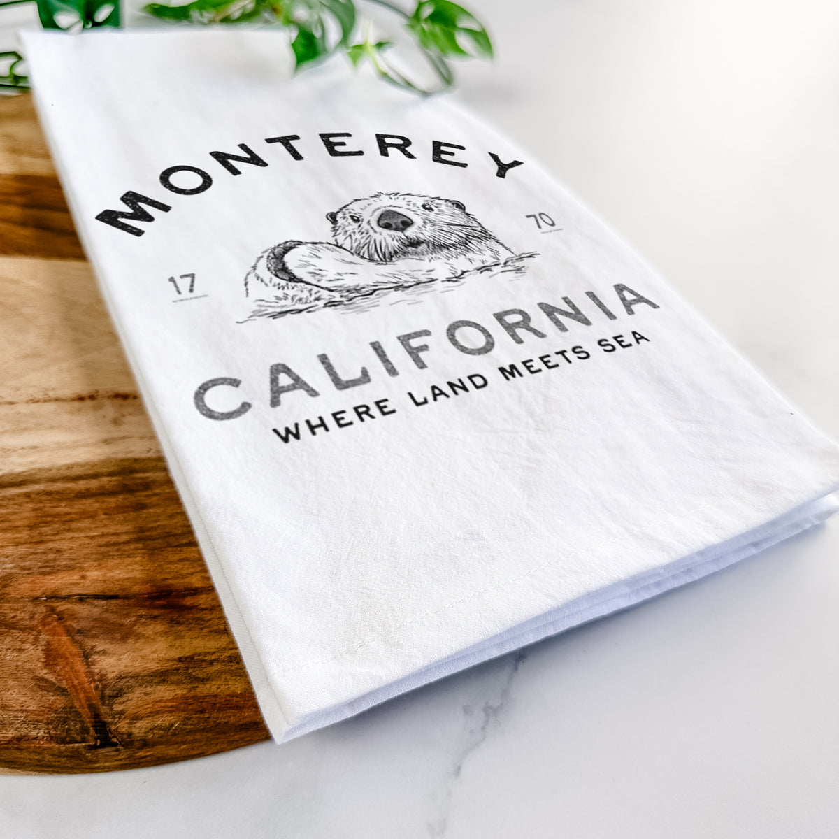 Close-up of a folded white tea towel on a wooden board, showing a black ink sea otter floating on its back, with the words Monterey California, 1770, Where Land Meets Sea.
