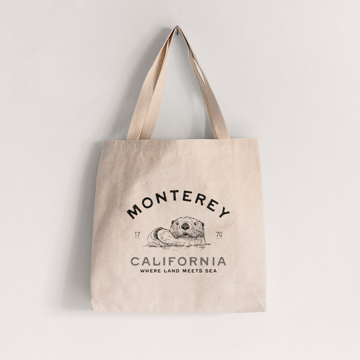Natural canvas tote bag printed with a black ink sea otter floating on its back, with the words Monterey California, 1770, Where Land Meets Sea.