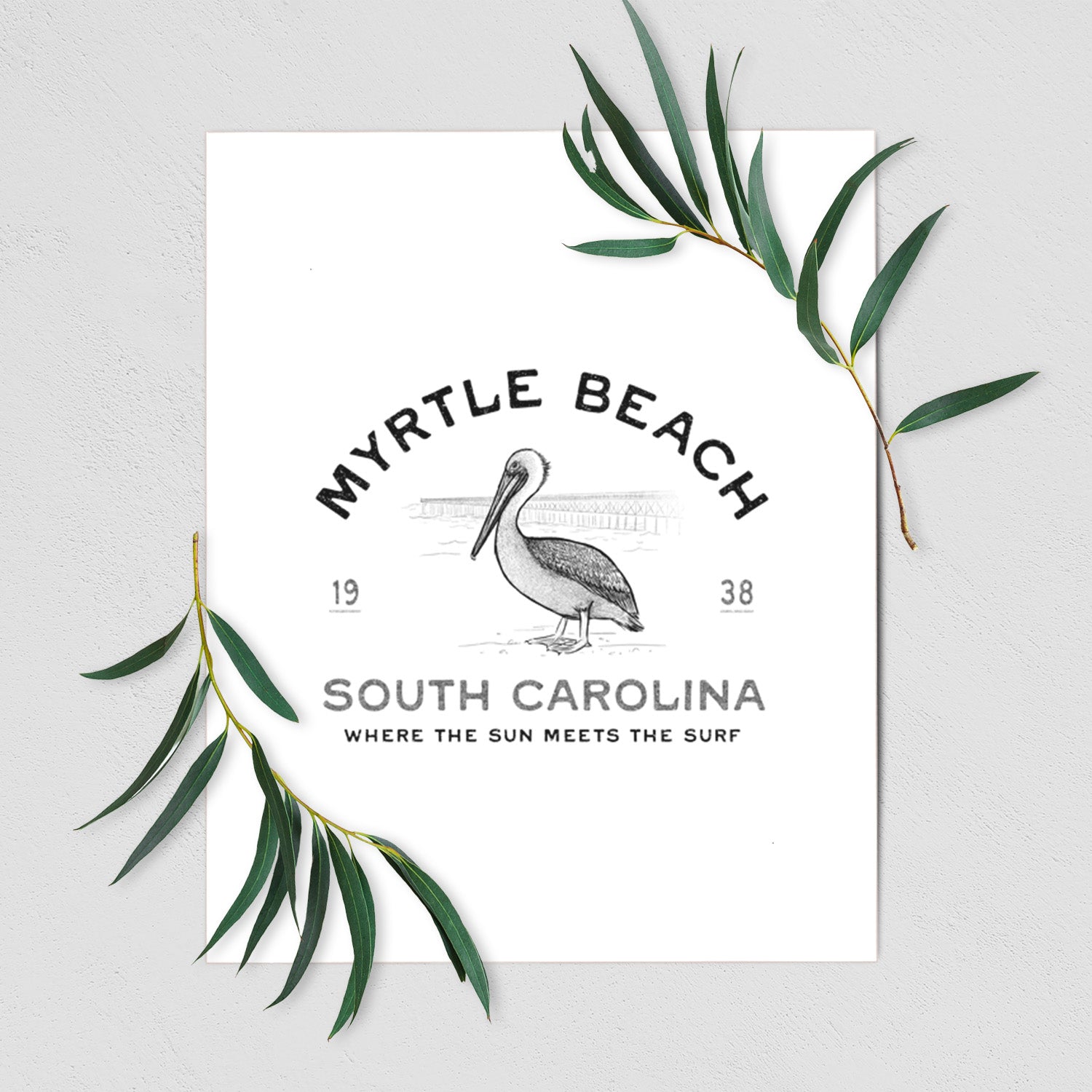 Art print of a black ink pelican on a post with the words Myrtle Beach South Carolina, Where the Sun Meets the Surf, shown with eucalyptus leaves.
