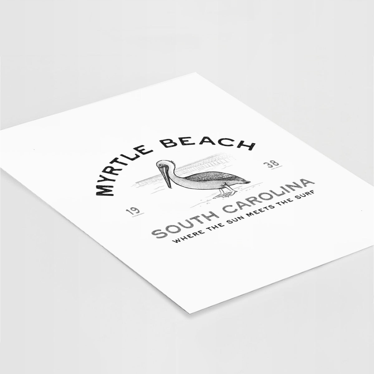 Art print of a black ink pelican on a post with the words Myrtle Beach South Carolina, Where the Sun Meets the Surf, lying at an angle on a white surface.