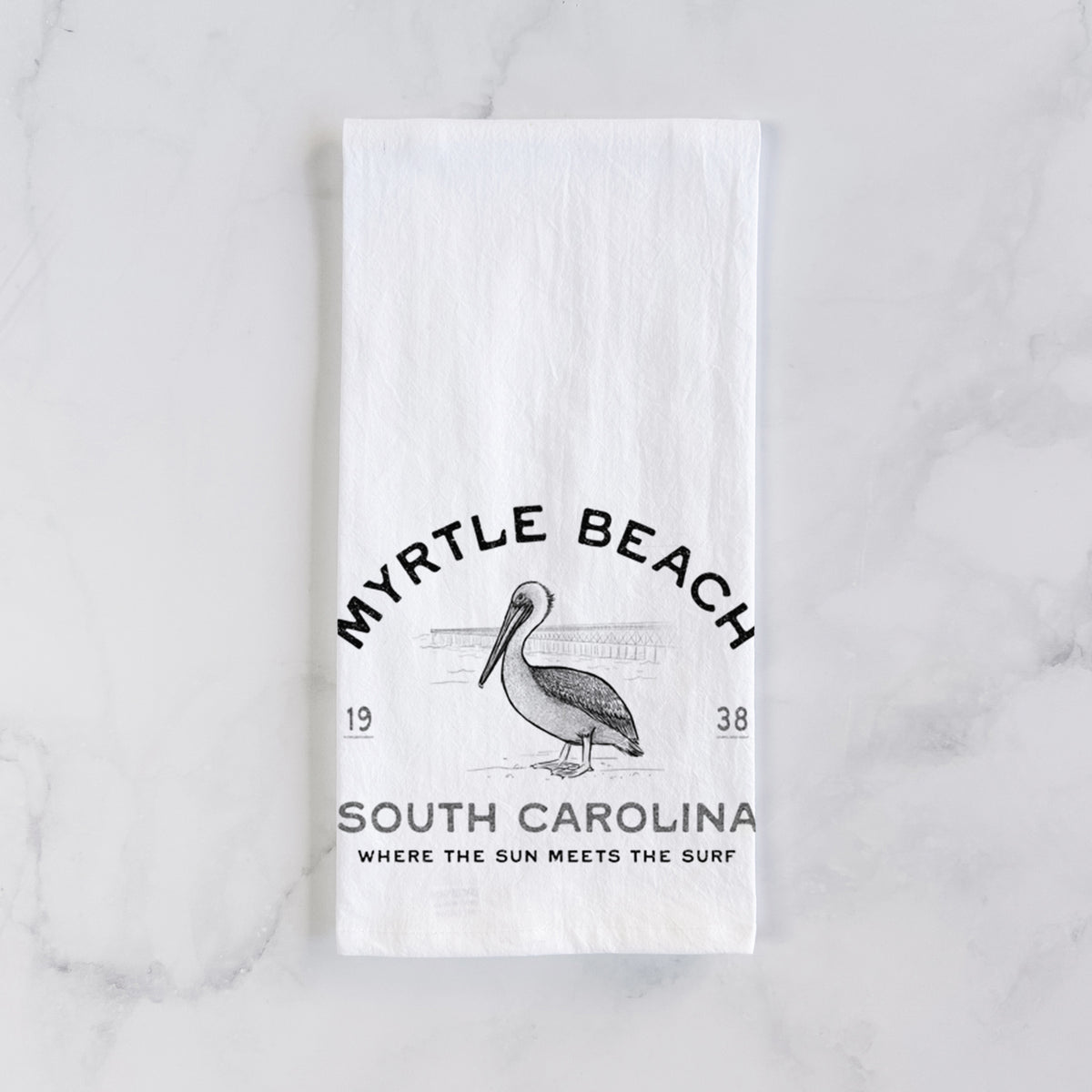 White cotton tea towel laid flat on marble, printed with a black ink pelican on a post with the words Myrtle Beach South Carolina, Where the Sun Meets the Surf.