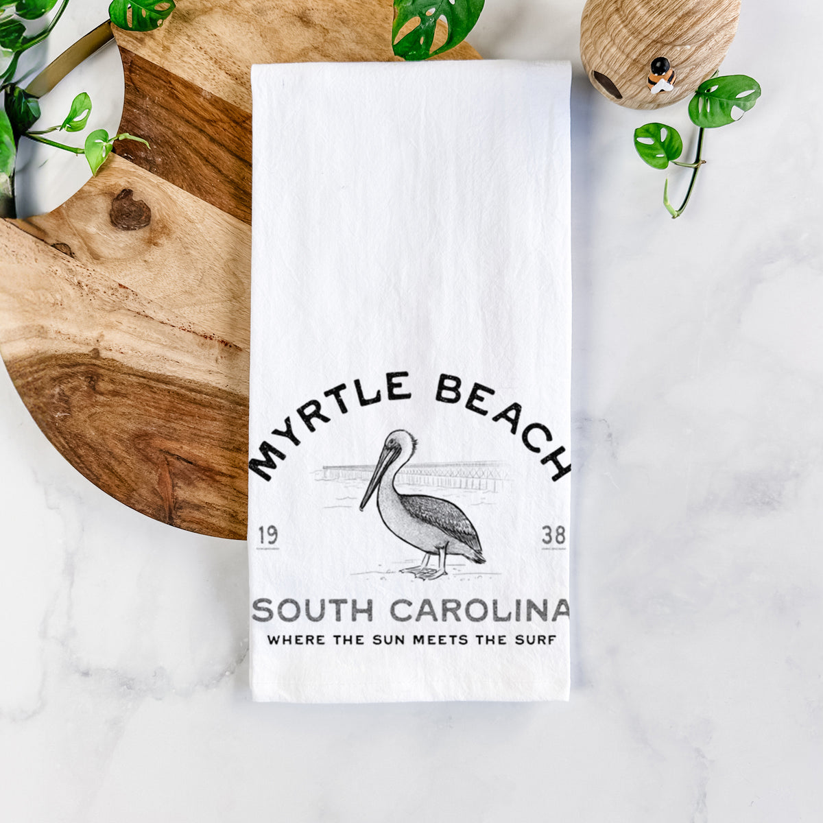White cotton tea towel printed with a black ink pelican on a post with the words Myrtle Beach South Carolina, Where the Sun Meets the Surf, on a marble counter beside a round wooden cutting board and greenery.