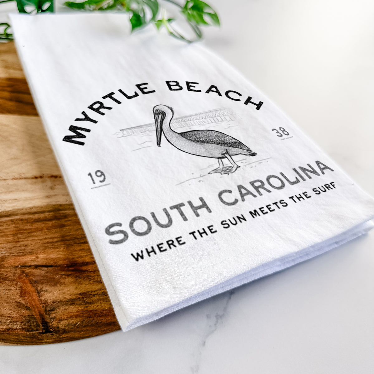 Close-up of a folded white tea towel on a wooden board, showing a black ink pelican on a post with the words Myrtle Beach South Carolina, Where the Sun Meets the Surf.