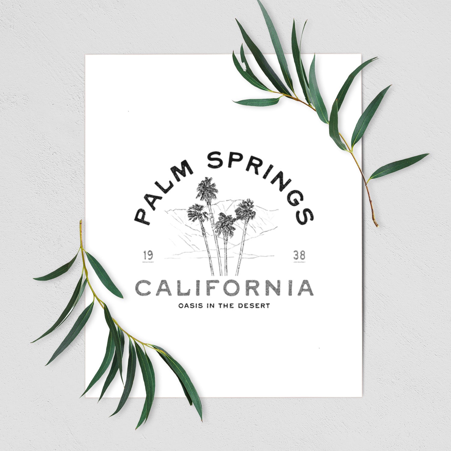 Art print of black ink palm trees with the words Palm Springs California, 1938, Oasis in the Desert, shown with eucalyptus leaves.