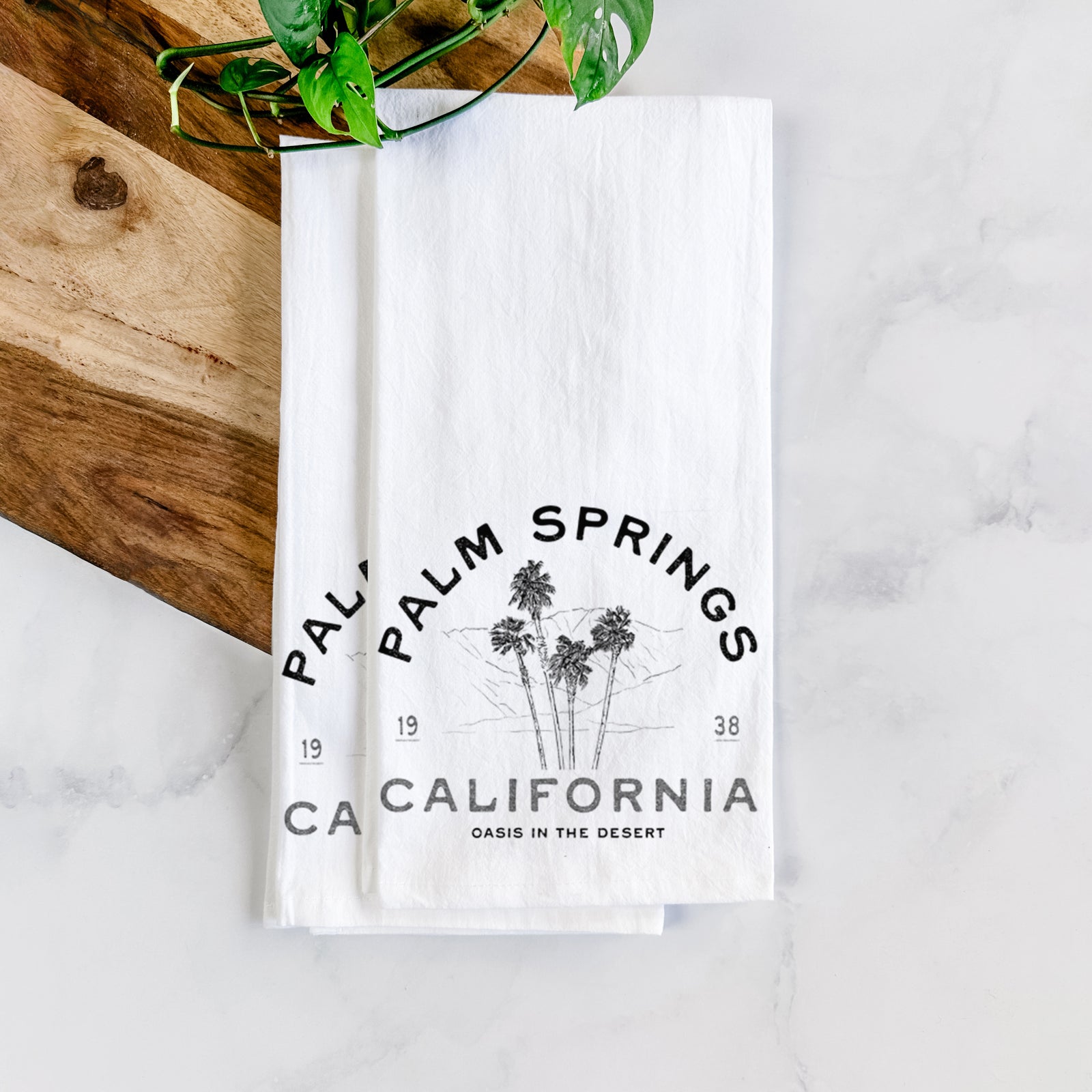 White cotton tea towel printed with black ink palm trees with the words Palm Springs California, 1938, Oasis in the Desert, on a marble counter with a wooden cutting board and greenery.
