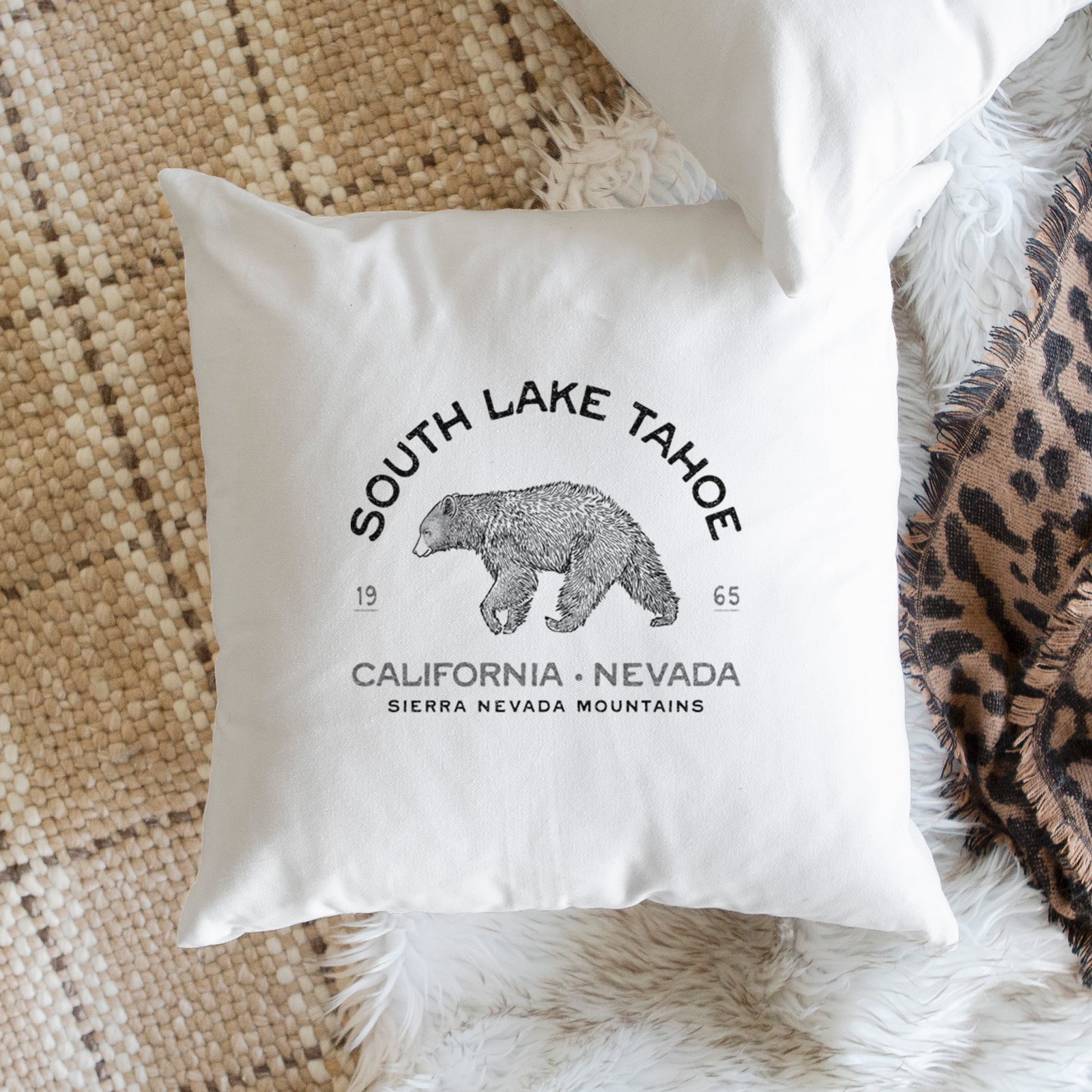White pillow cover printed with a black ink bear with the words South Lake Tahoe, California - Nevada, Sierra Nevada Mountains, on a bench beside plants.