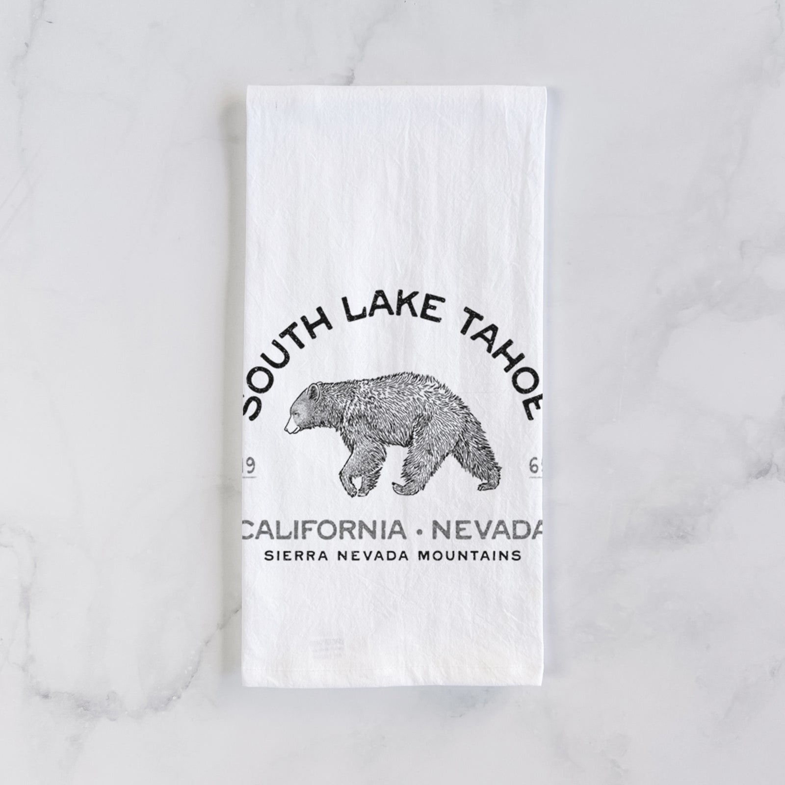 White cotton tea towel printed with a black ink bear with the words South Lake Tahoe, California - Nevada, Sierra Nevada Mountains, on a marble counter beside a round wooden cutting board and greenery.