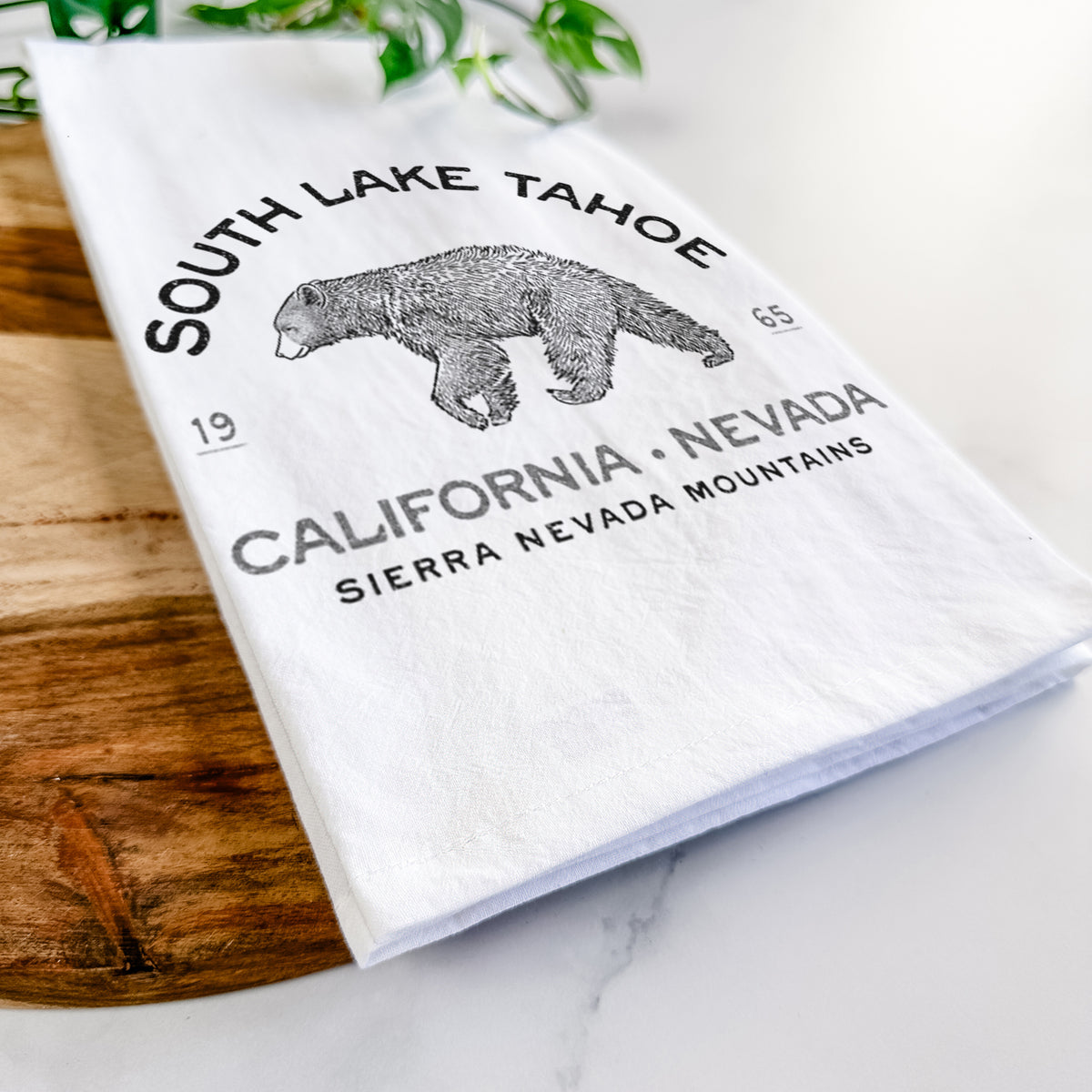Close-up of a folded white tea towel on a wooden board, showing a black ink bear with the words South Lake Tahoe, California - Nevada, Sierra Nevada Mountains.