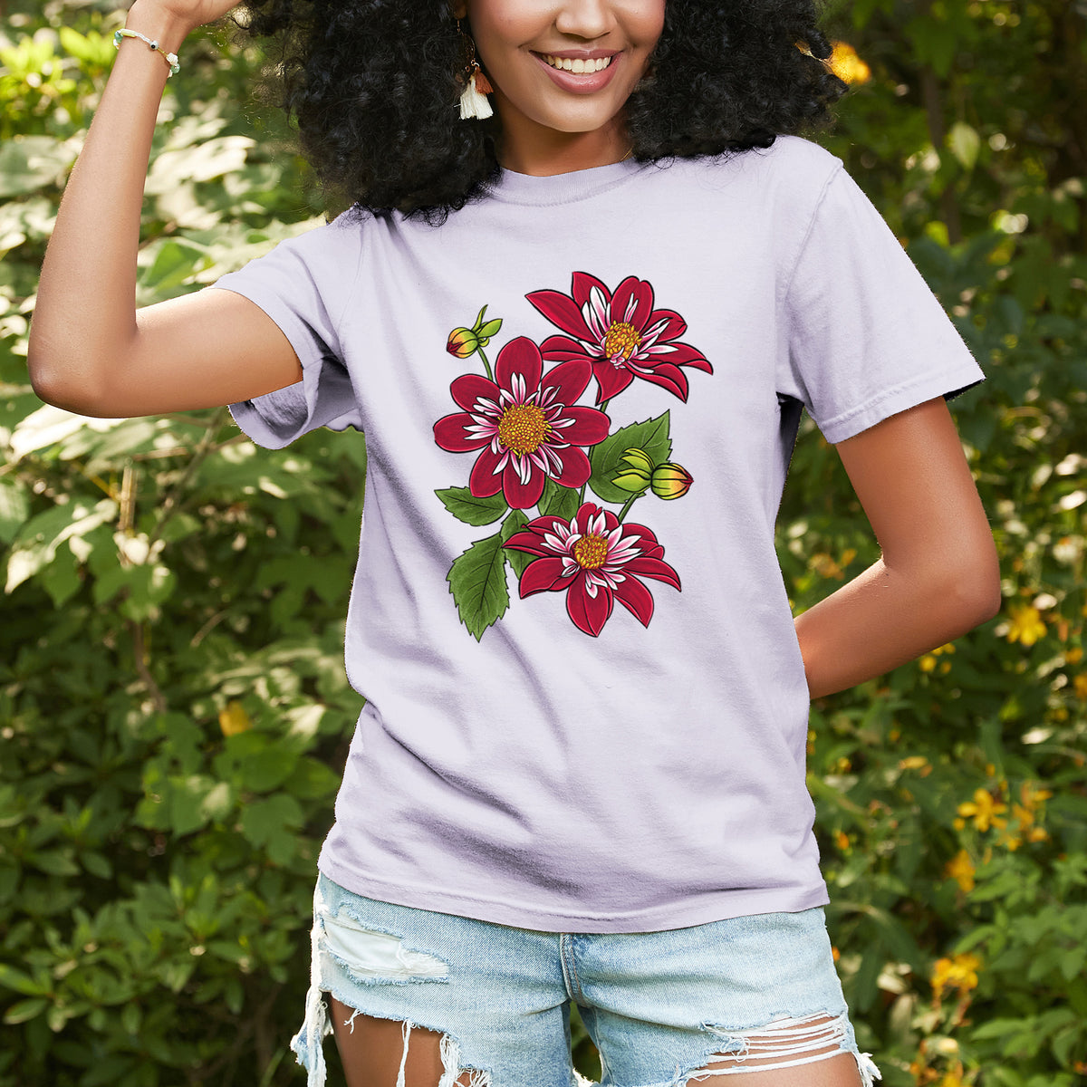 Woman wearing a Comfort Colors t-shirt printed with a full-color illustration of deep red dahlia flowers with green leaves and a small bee, outdoors.
