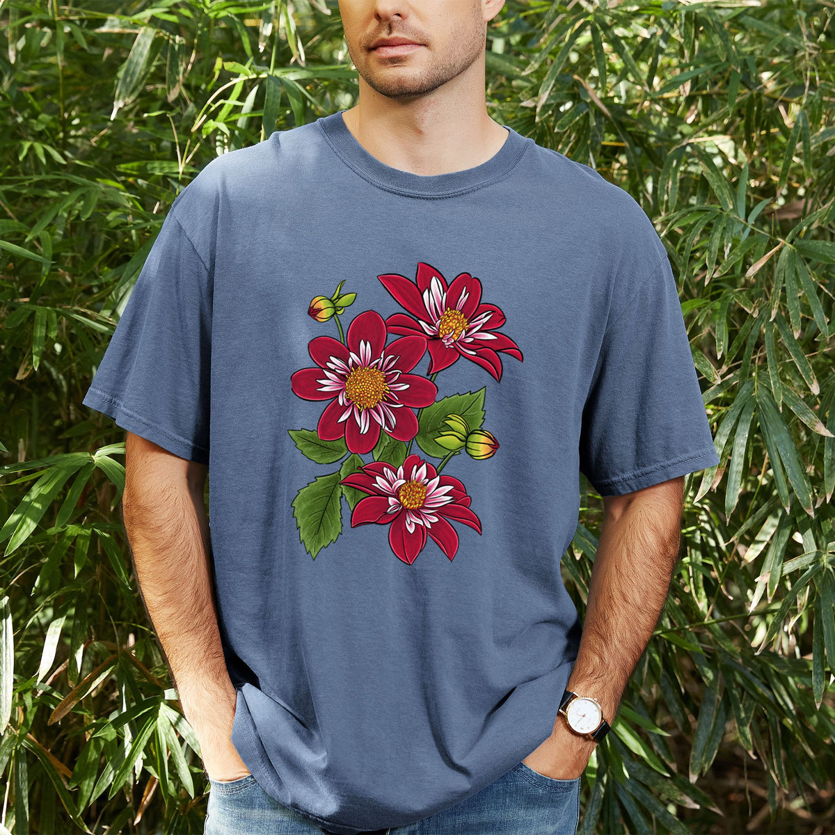 Man wearing a Comfort Colors t-shirt printed with a full-color illustration of deep red dahlia flowers with green leaves and a small bee, outdoors.