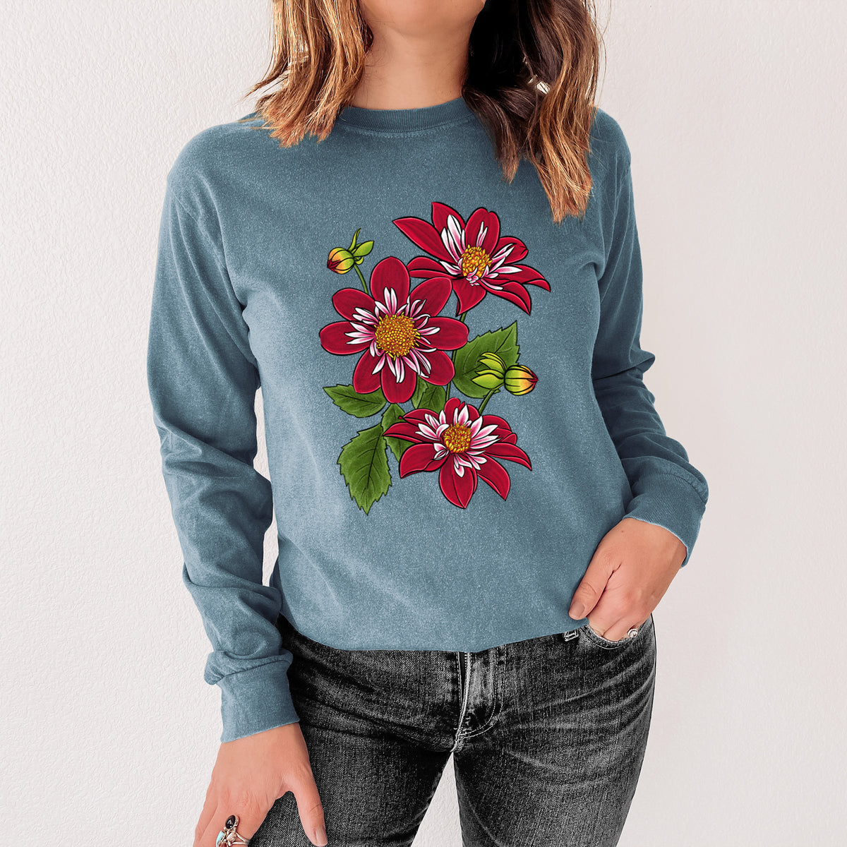 Steel blue Comfort Colors long sleeve shirt printed with a full-color illustration of deep red dahlia flowers with green leaves and a small bee, shown in a lifestyle photo.