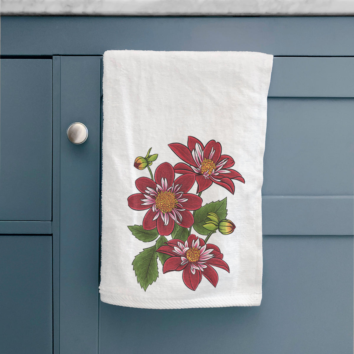 White hand towel printed with a full-color illustration of deep red dahlia flowers with green leaves and a small bee, hanging on a cabinet door.