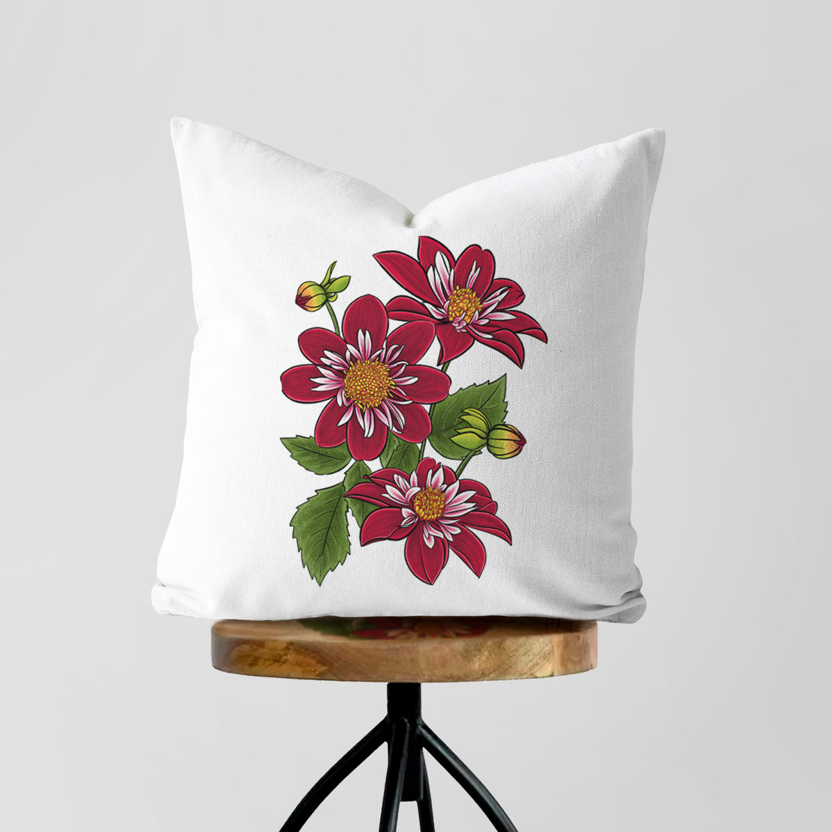 White pillow cover printed with a full-color illustration of deep red dahlia flowers with green leaves and a small bee, on a wooden stool.