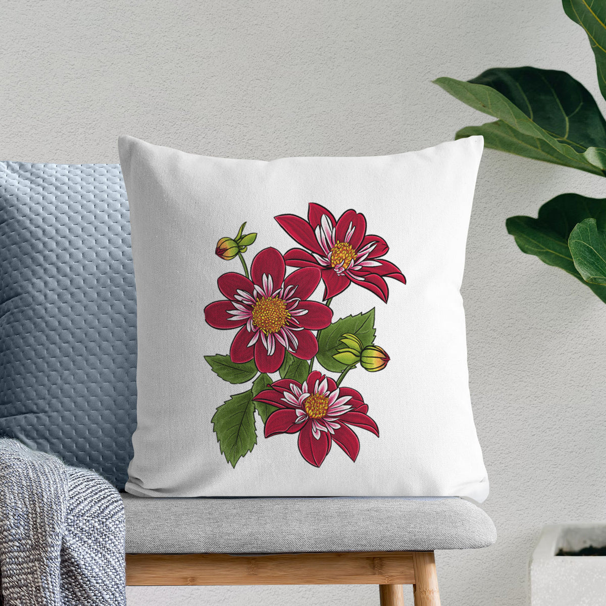 White pillow cover printed with a full-color illustration of deep red dahlia flowers with green leaves and a small bee, on a bench beside plants.