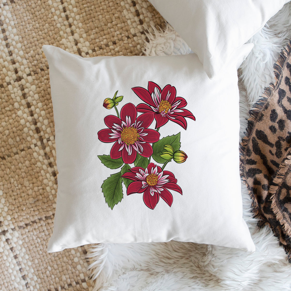 White pillow cover printed with a full-color illustration of deep red dahlia flowers with green leaves and a small bee, on a woven chair.