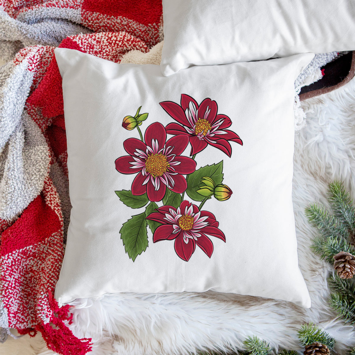 White pillow cover printed with a full-color illustration of deep red dahlia flowers with green leaves and a small bee, on a woven chair.