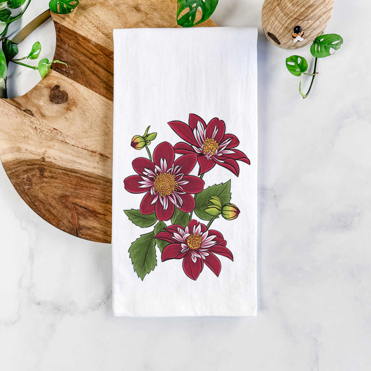 White cotton tea towel printed with a full-color illustration of deep red dahlia flowers with green leaves and a small bee, on a marble counter beside a round wooden cutting board and greenery.
