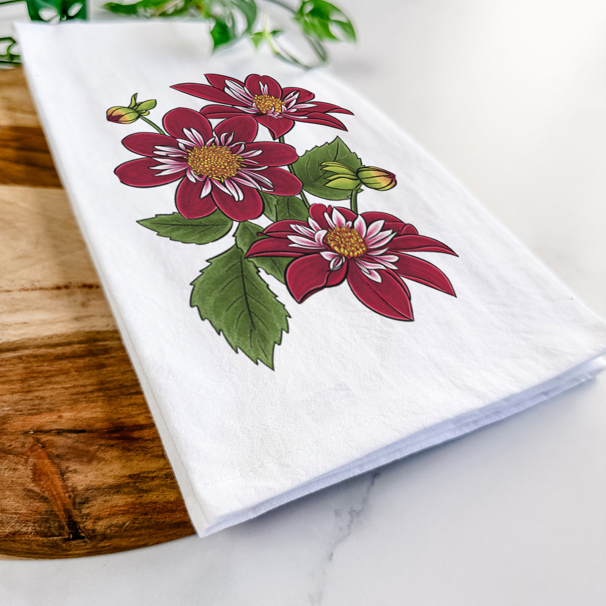 Close-up of a folded white tea towel on a wooden board, showing a full-color illustration of deep red dahlia flowers with green leaves and a small bee.