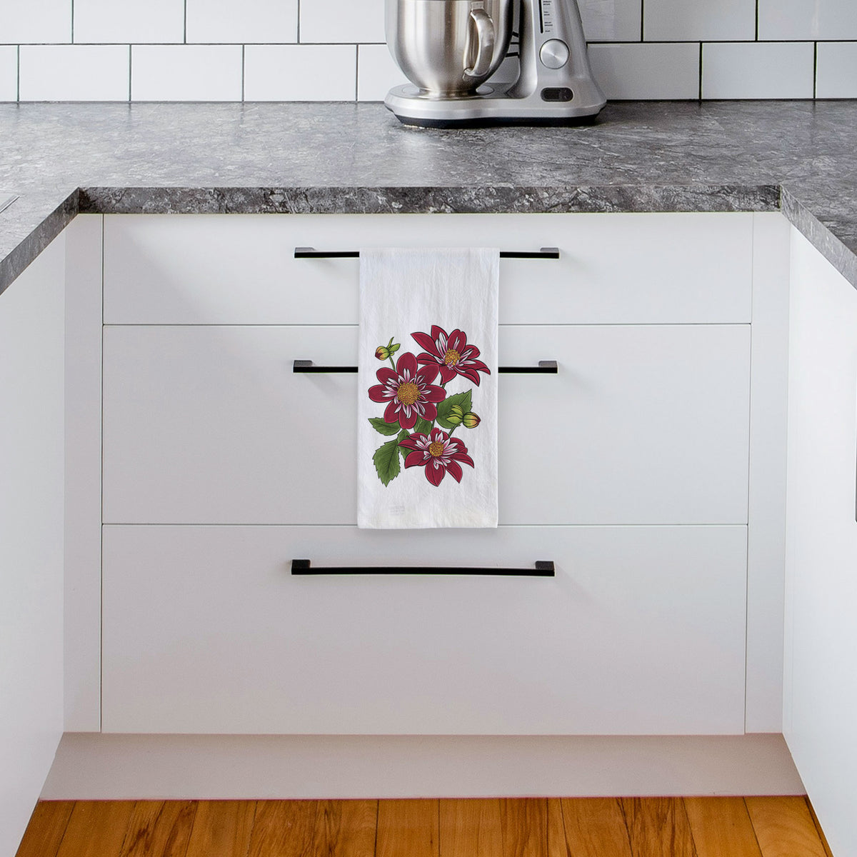 White tea towel printed with a full-color illustration of deep red dahlia flowers with green leaves and a small bee, hanging from a drawer handle in a bright white kitchen.
