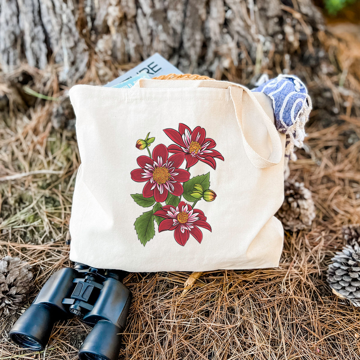 Canvas tote bag printed with a full-color illustration of deep red dahlia flowers with green leaves and a small bee, resting on dry grass beside a pair of binoculars.