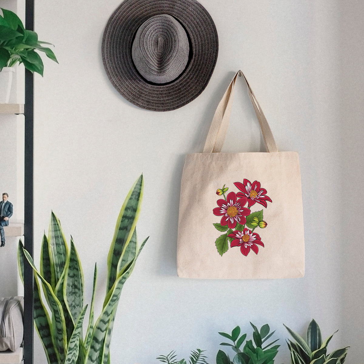 Canvas tote bag printed with a full-color illustration of deep red dahlia flowers with green leaves and a small bee, hanging on a wall hook below a hat, next to potted plants.