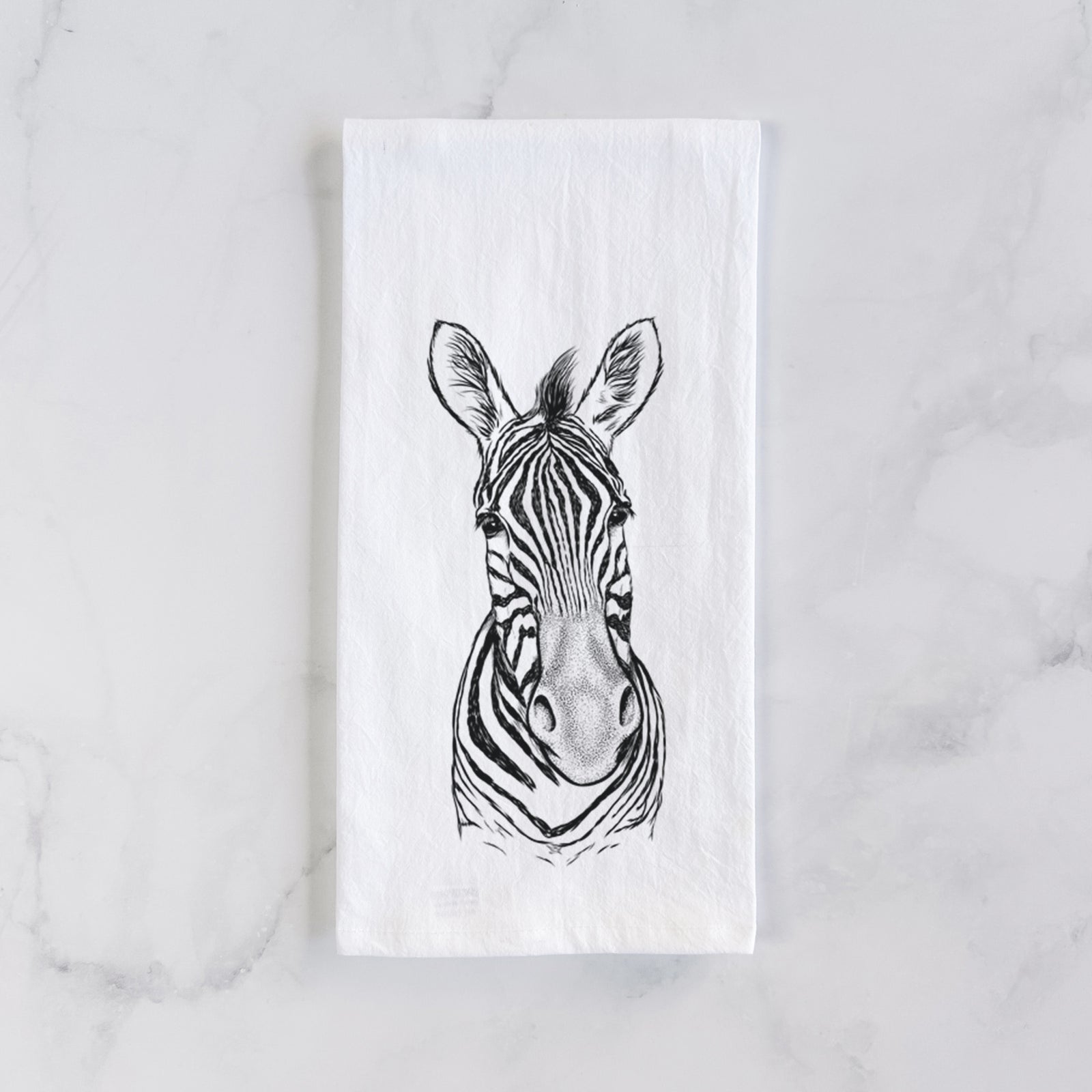White cotton tea towel printed with a detailed black ink portrait of a Damara zebra facing forward, on a marble counter beside a round wooden cutting board and greenery.