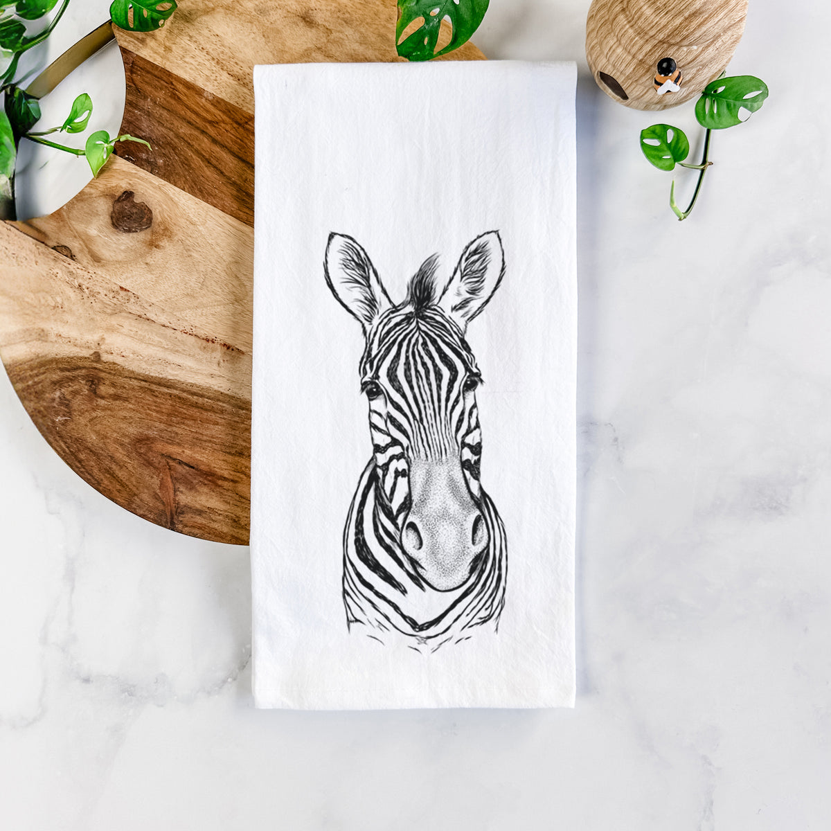 White cotton tea towel printed with a detailed black ink portrait of a Damara zebra facing forward, on a marble counter beside a round wooden cutting board and greenery.