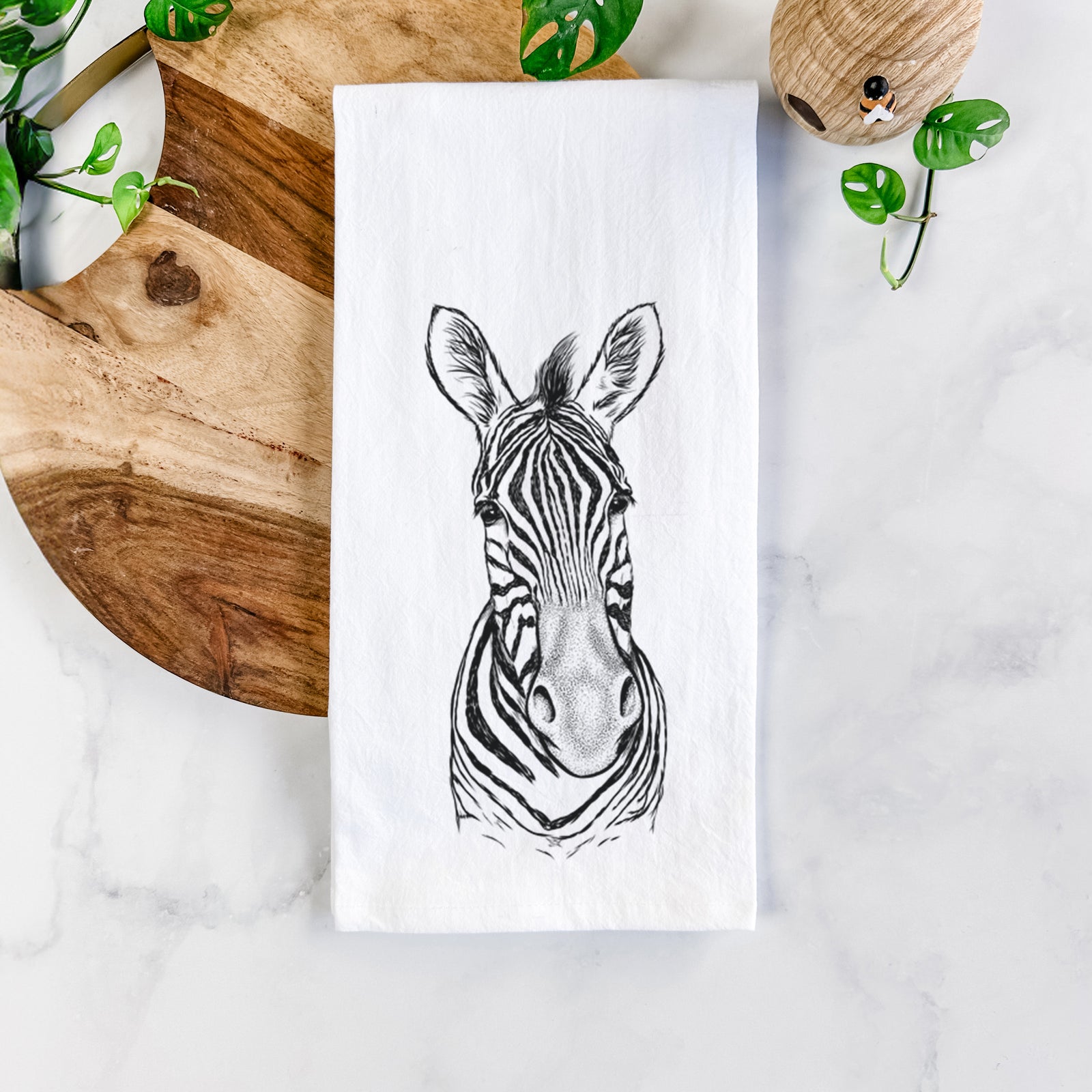 White cotton tea towel printed with a detailed black ink portrait of a Damara zebra facing forward, on a marble counter beside a round wooden cutting board and greenery.