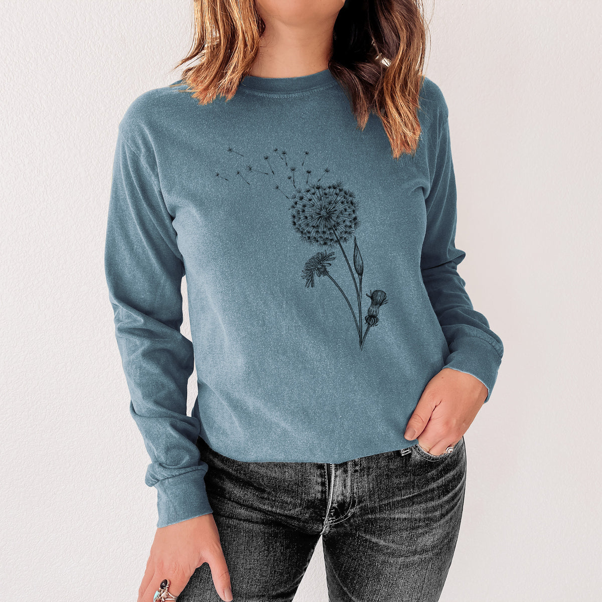 Steel blue Comfort Colors long sleeve shirt printed with a black ink line drawing of a dandelion seed head with seeds blowing away, shown in a lifestyle photo.