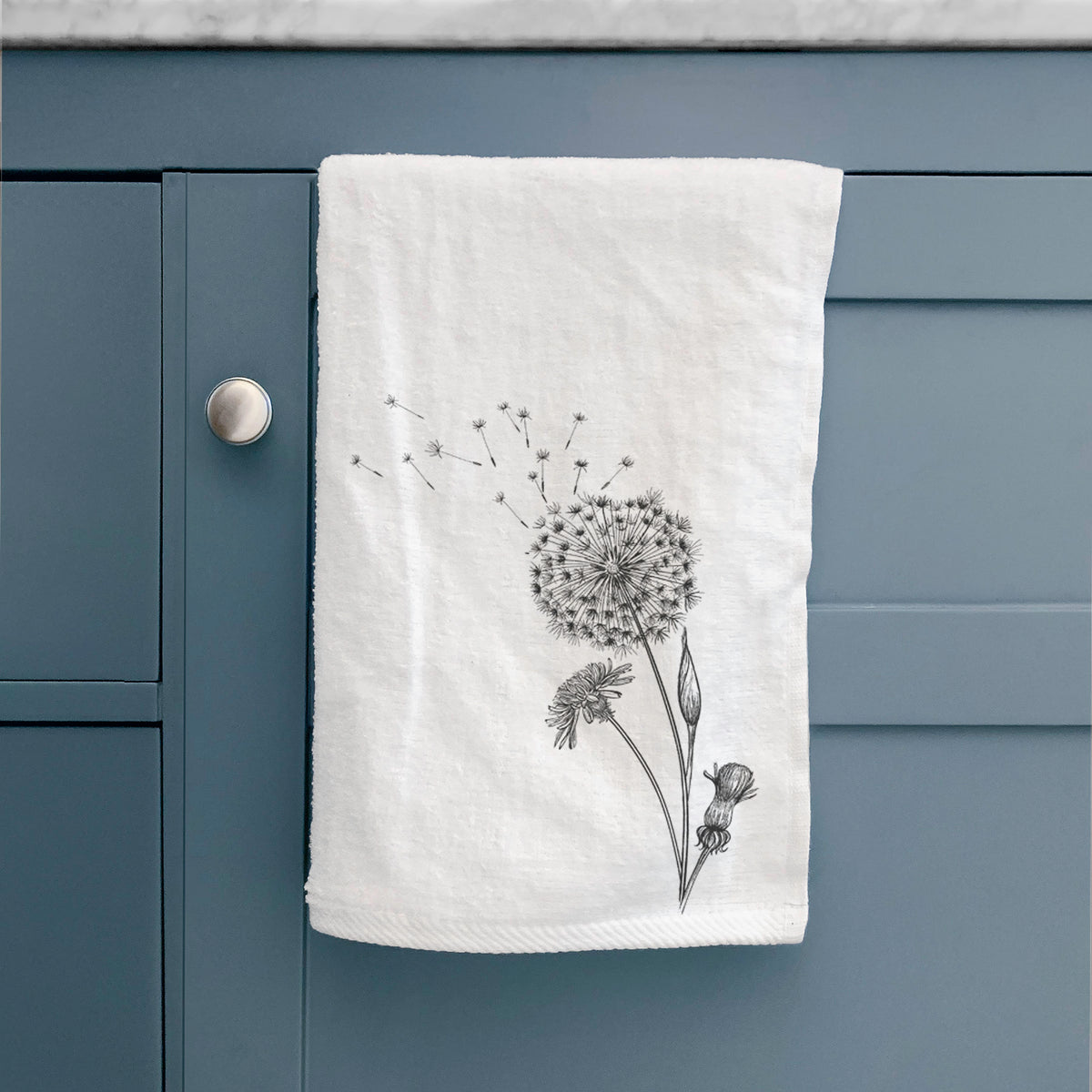 White hand towel printed with a black ink line drawing of a dandelion seed head with seeds blowing away, hanging on a cabinet door.