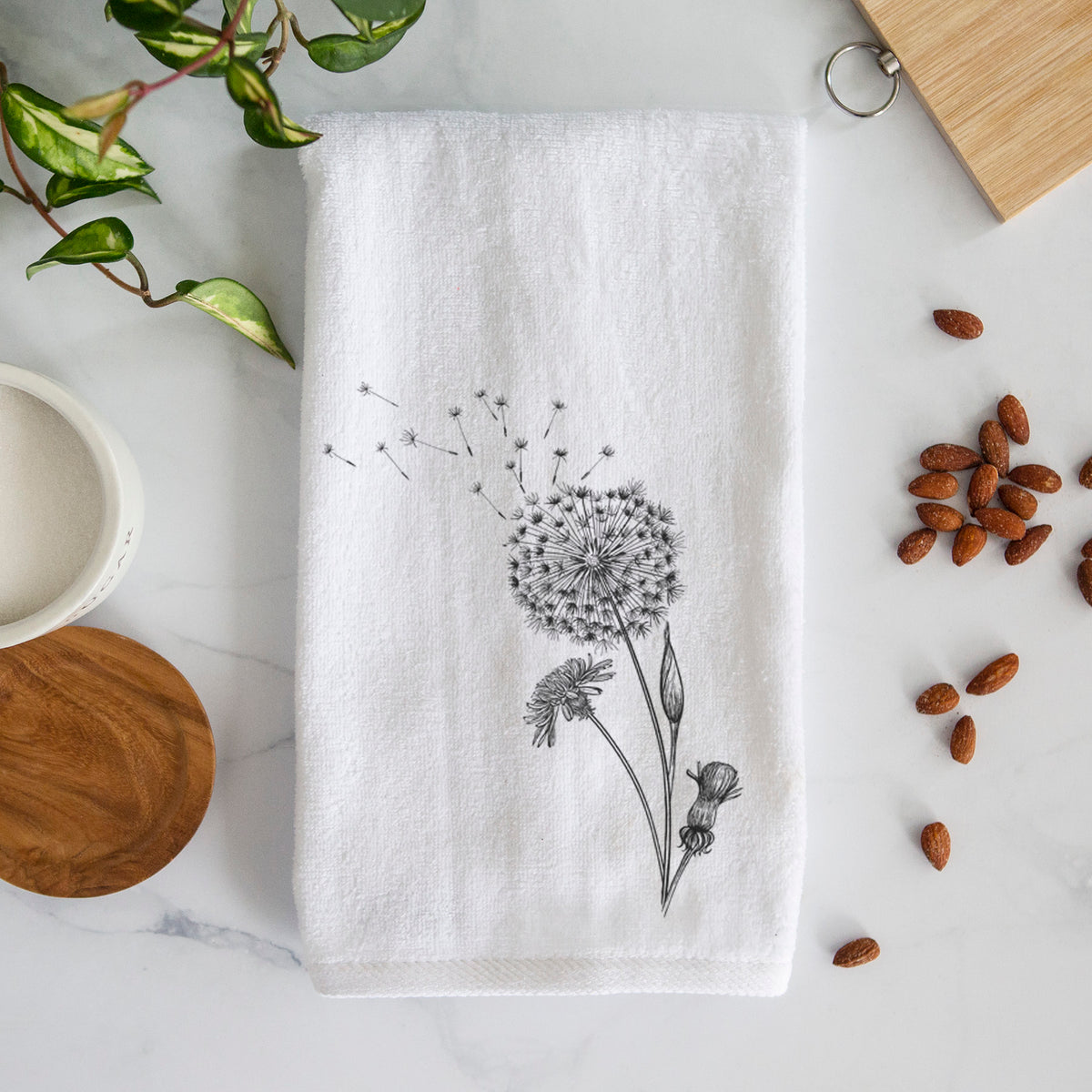 White hand towel printed with a black ink line drawing of a dandelion seed head with seeds blowing away.