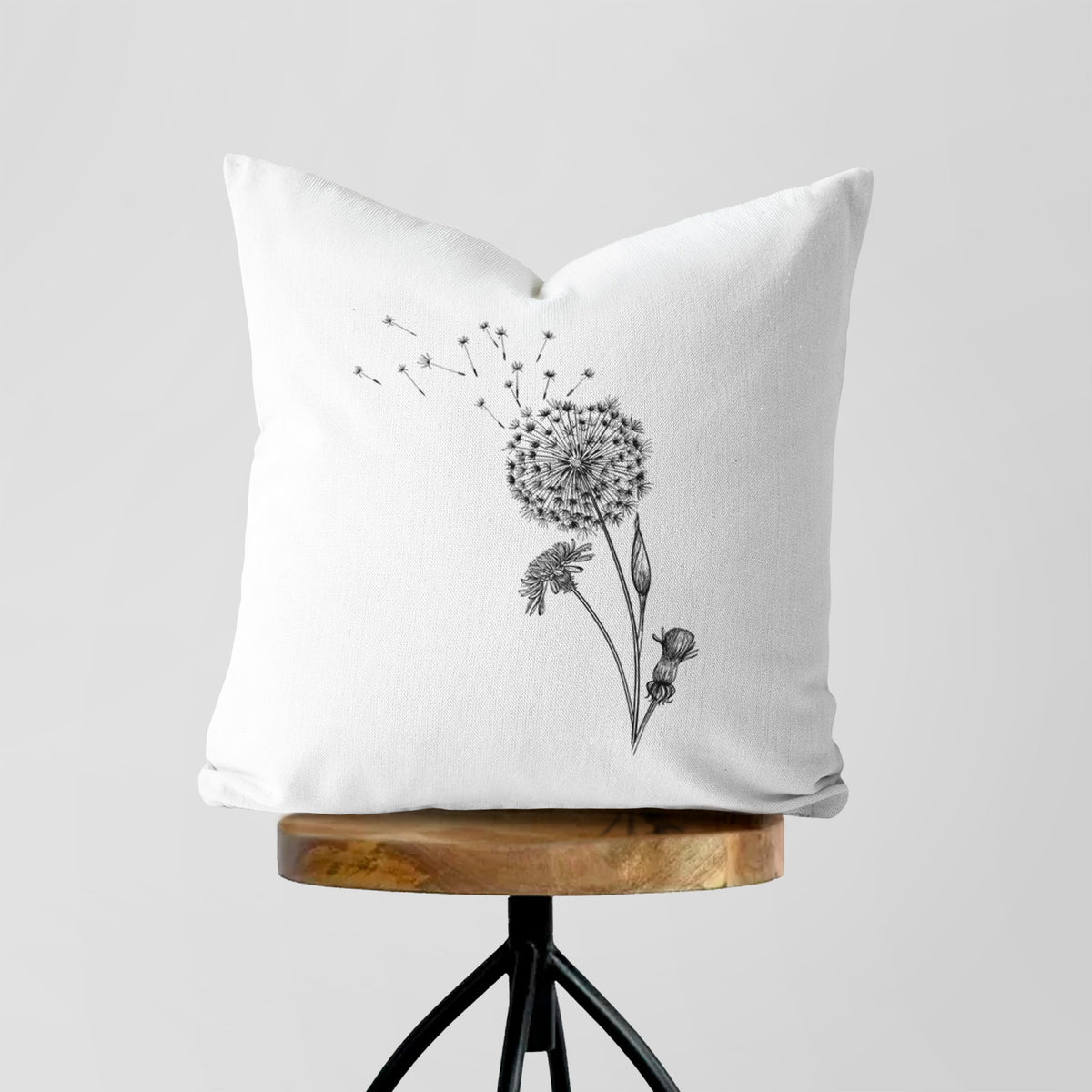 White pillow cover printed with a black ink line drawing of a dandelion seed head with seeds blowing away, on a wooden stool.