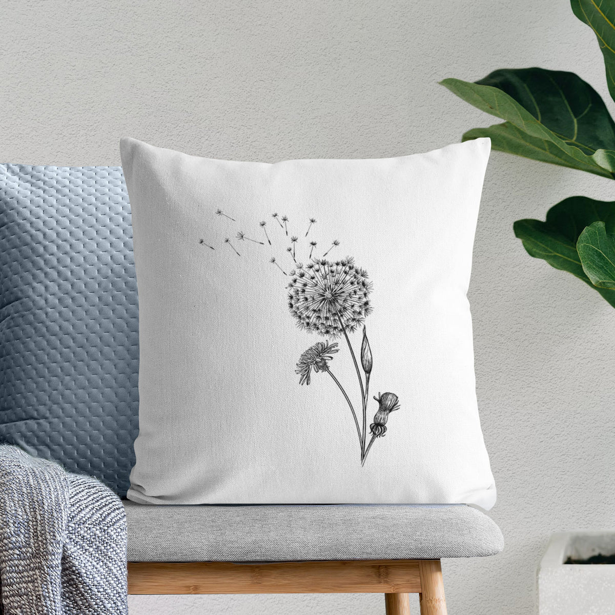White pillow cover printed with a black ink line drawing of a dandelion seed head with seeds blowing away, on a bench beside plants.